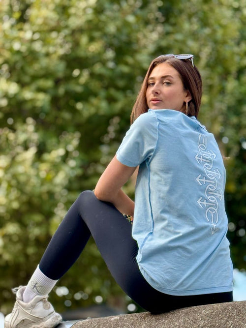 Woman sitting outdoors wearing a light blue t-shirt with text and dark leggings.
