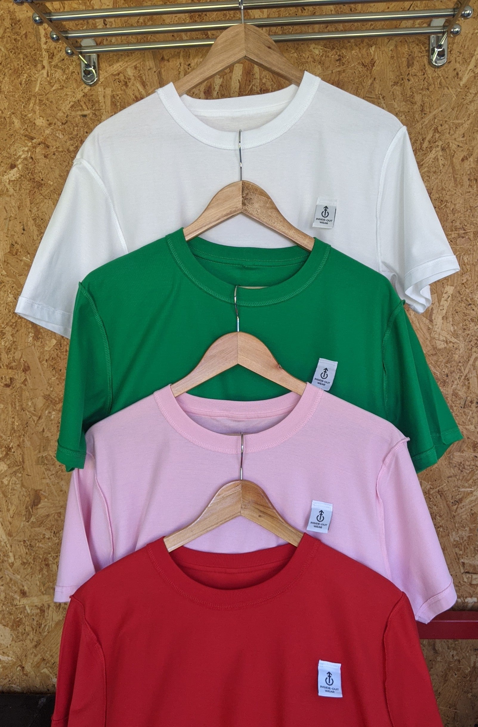 Four shirts in white, green, pink, and red hanging on wooden hangers against a corkboard background.